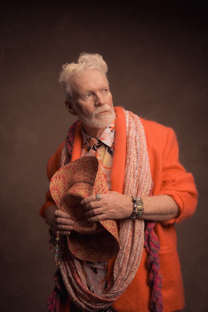 Stylish elderly man in vibrant orange jacket with colorful accessories, posed in studio setting.