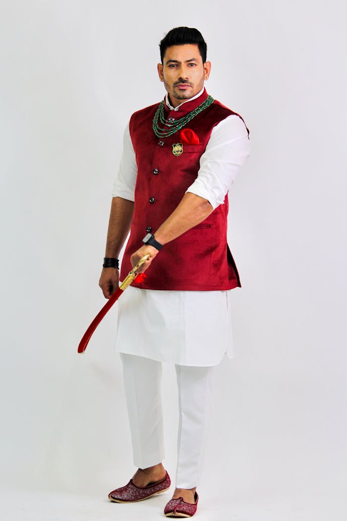 Indian man in traditional red attire holding a sword against a white studio background.